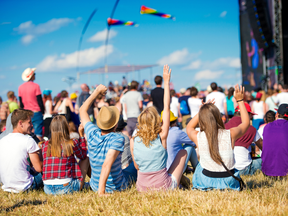 A group of friends sitting on the grass at a sunny music festival, with backs to the camera