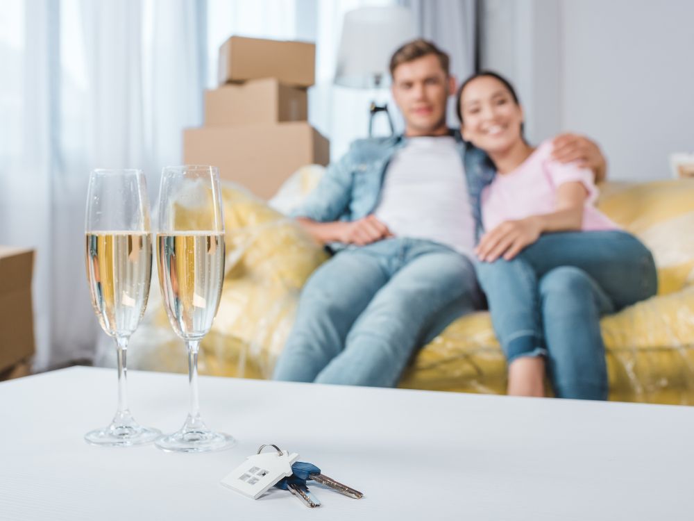 A couple relax on a couch in their new home, with keys and glasses of Champagne in the forreground